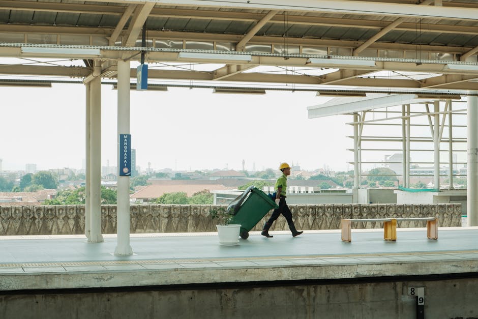 A person wearing a gray jacket and gray cap is standing outdoors near an entrance to a train platform, pushing a red trolley with a blue vacuum cleaner attached. The scene includes a concrete ground w