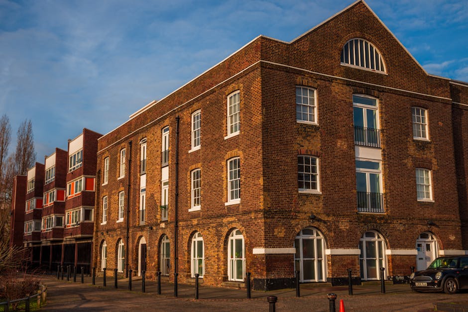 Exterior view of a multi-story brick residential building with arched and rectangular windows, some with balconies, under a partly cloudy sky during daylight. The building's façade shows signs of weat